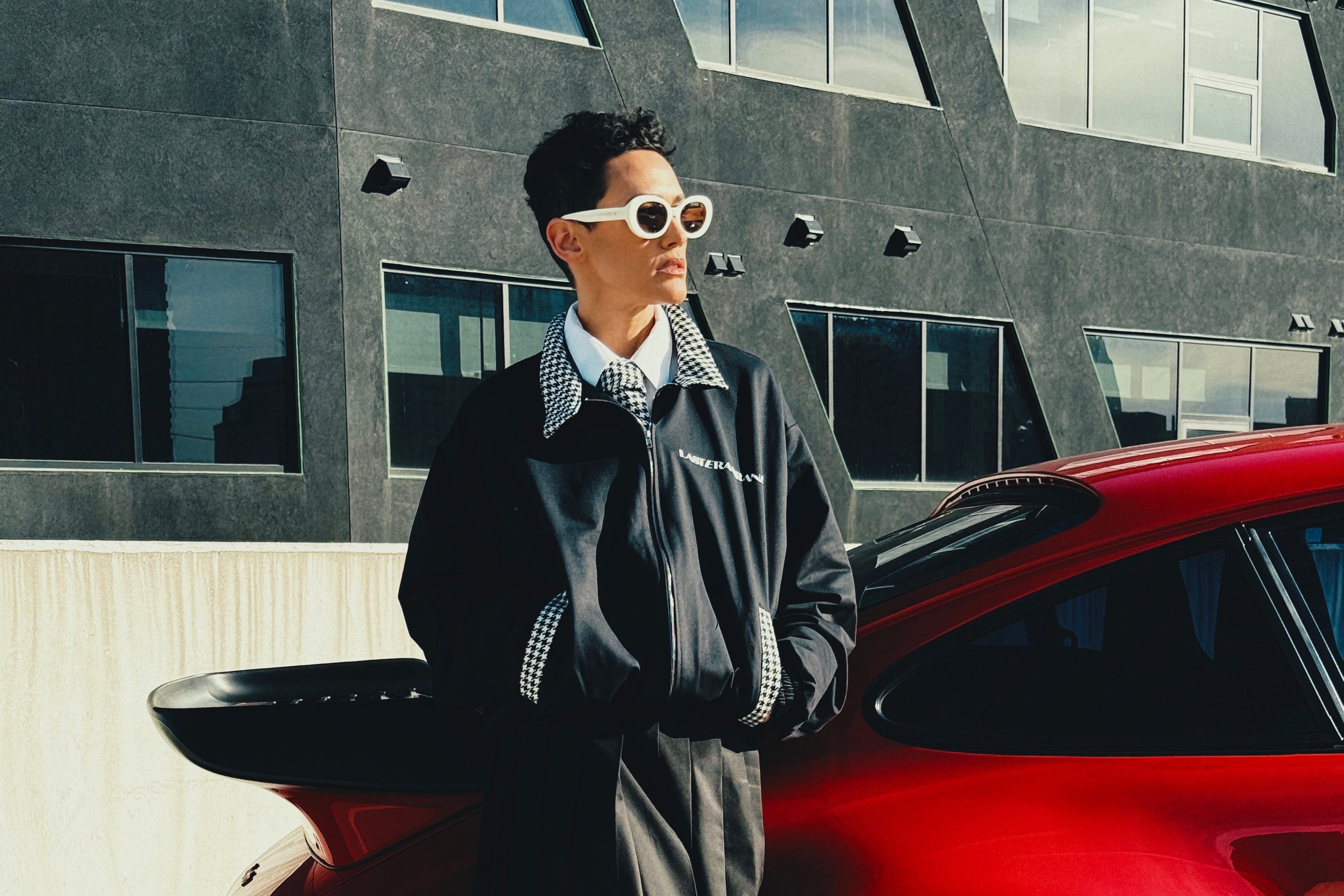 Person in a stylish outfit standing next to a red car with a modern building in the background